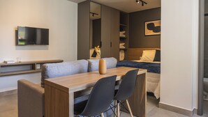 Une chambre d’hôtel moderne avec un bureau en bois, deux chaises noires, un grand miroir, un lit recouvert d’une couverture bleue et un téléviseur à écran plat fixé au mur.