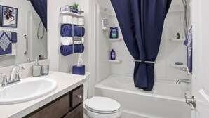 Et badeværelse med en hvid vask, toilet og badekar. Håndklæder og toiletartikler er placeret på hylder.