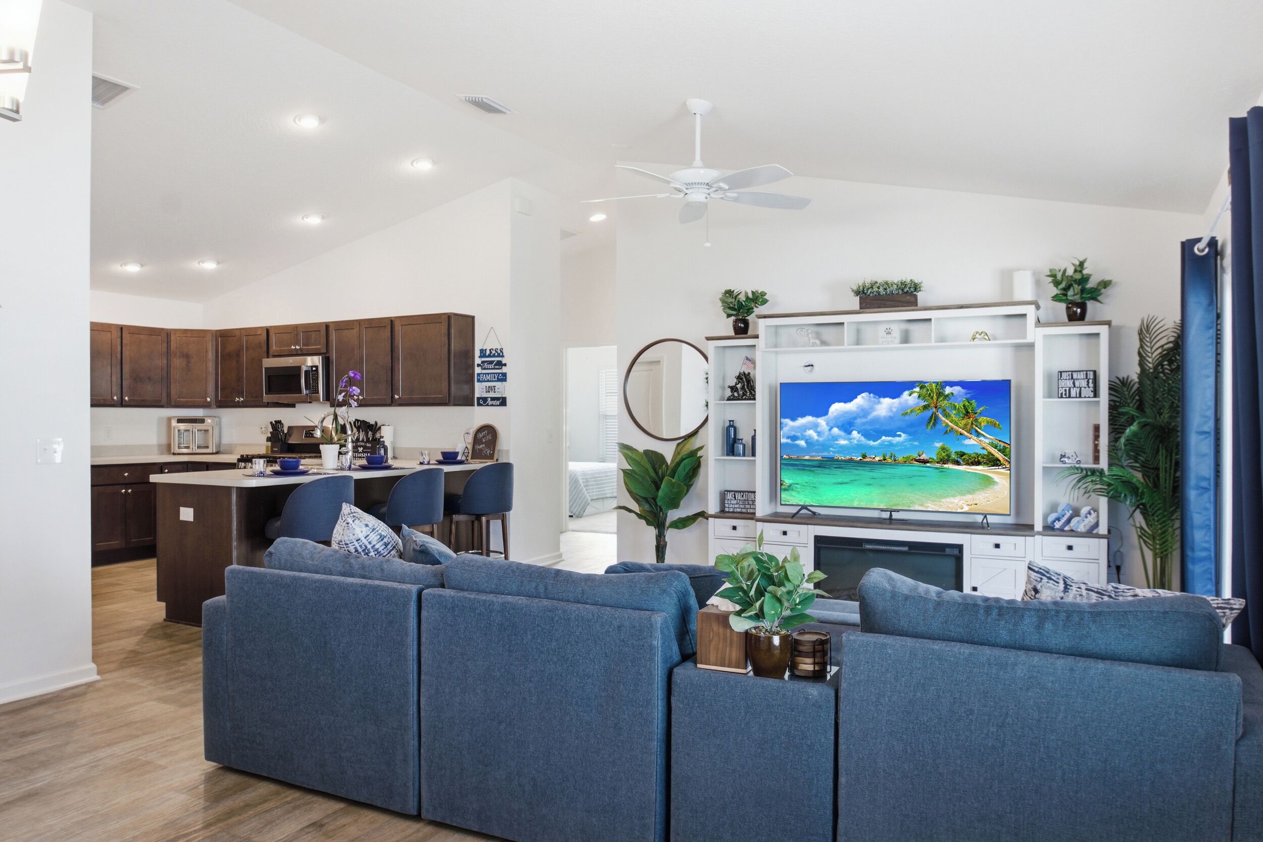 A modern living room with a large flat-screen TV, a sofa, and a dining area with a kitchen in the background.