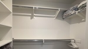 Ein begehbarer Kleiderschrank mit weißen Regalen, einer Kleiderstange und einem Bett mit weißer Bettwäsche.