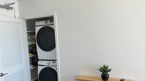Ein Waschraum mit zwei übereinander gestapelten Waschmaschinen, einem Holztischchen mit einer Pflanze und einer weißen Tür mit Lüftungsgitter darüber.