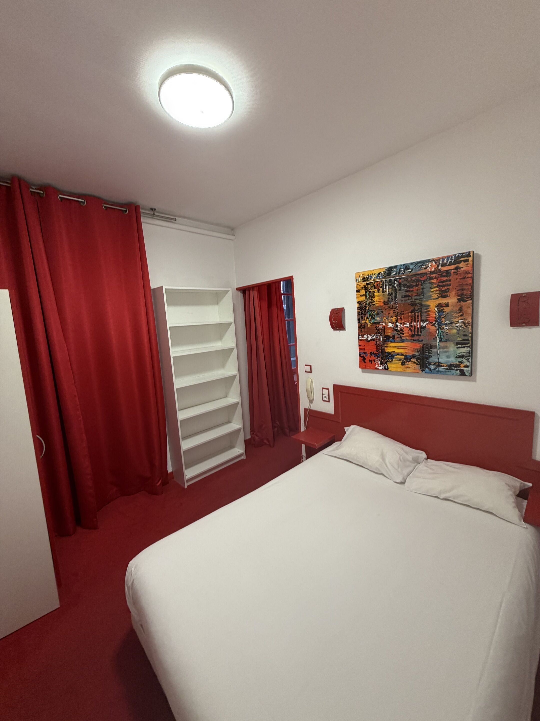 A hotel room with a bed, a white wardrobe, a red curtain, a red headboard, and a colorful painting on the wall.