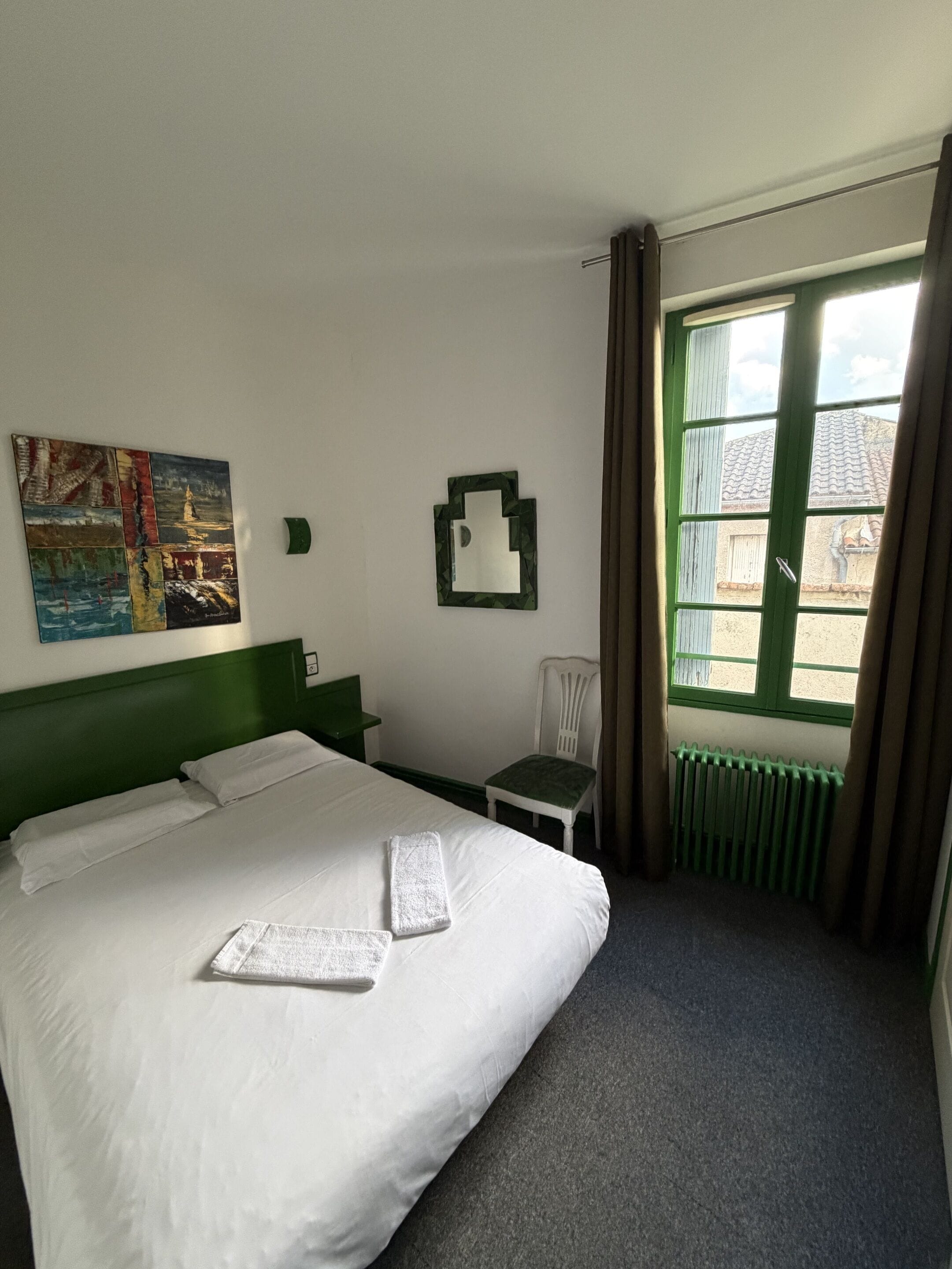 A hotel room with a large bed, a small chair, a radiator, and a window with curtains.