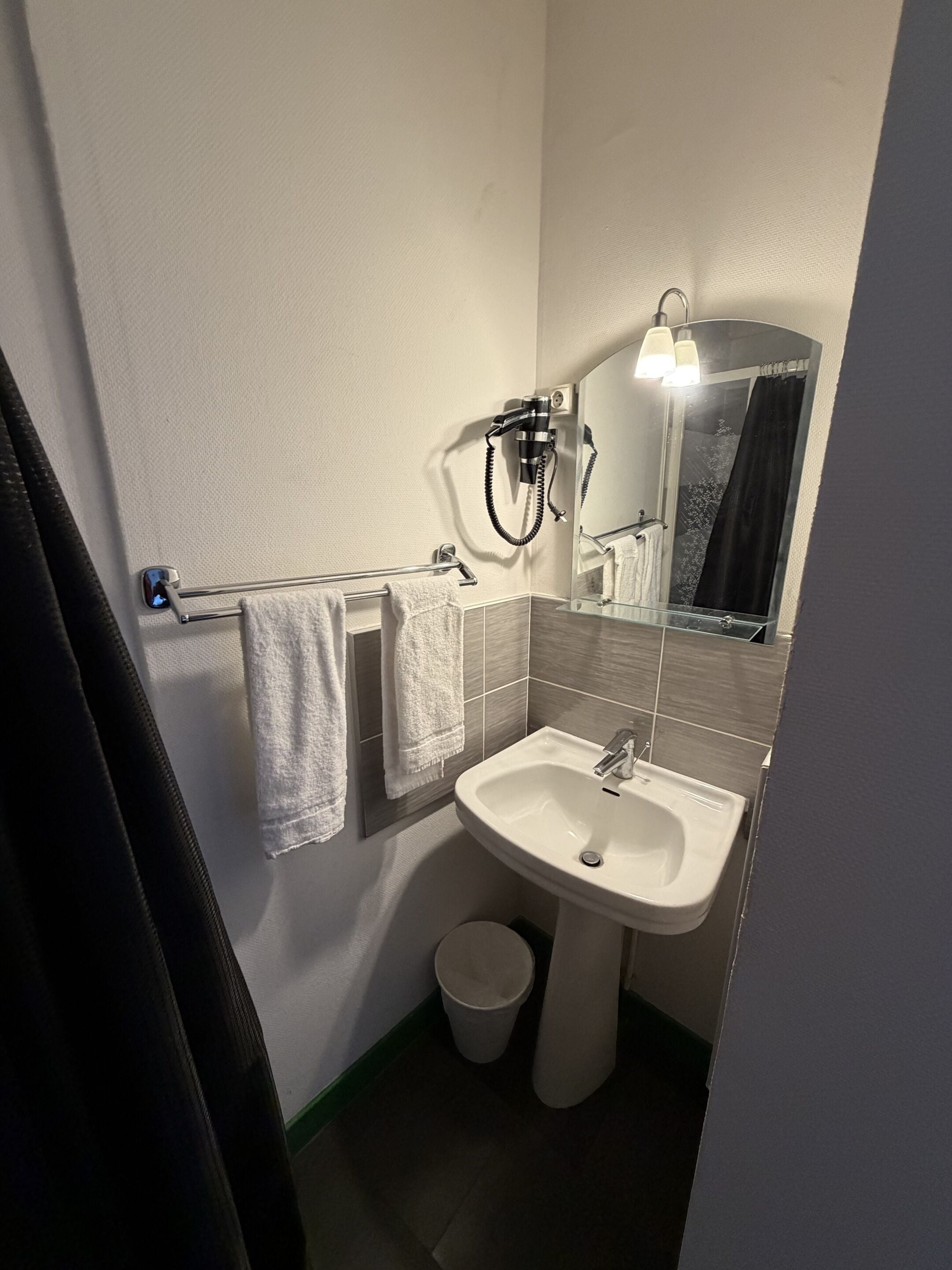 A compact bathroom with a white sink, a towel rack with two towels, a hairdryer, a mirror, and a small trash bin.