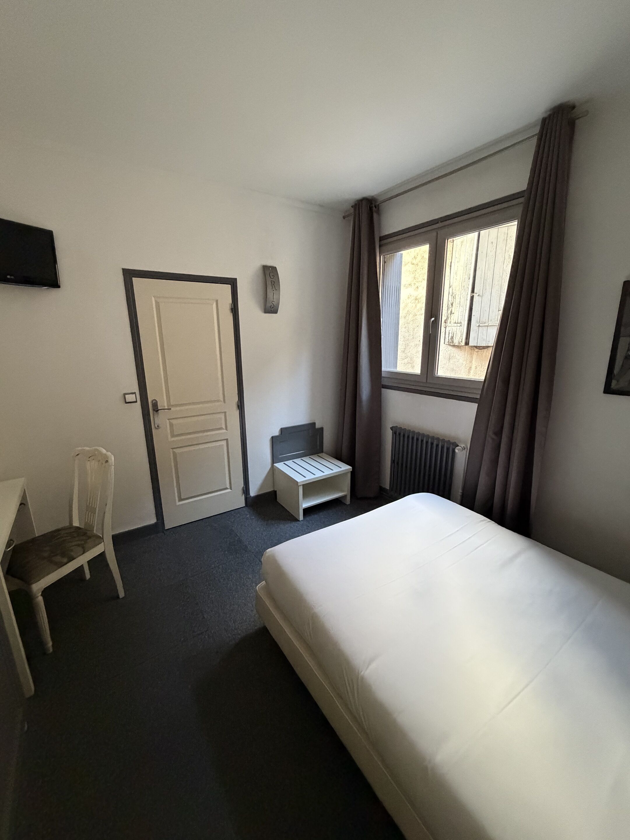 A hotel room with a bed, a chair, a small table, a window with curtains, and a wall-mounted TV.