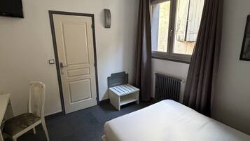 A hotel room with a bed, a chair, a small table, a window with curtains, and a wall-mounted TV.