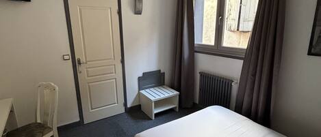 A hotel room with a bed, a chair, a small table, a window with curtains, and a wall-mounted TV.