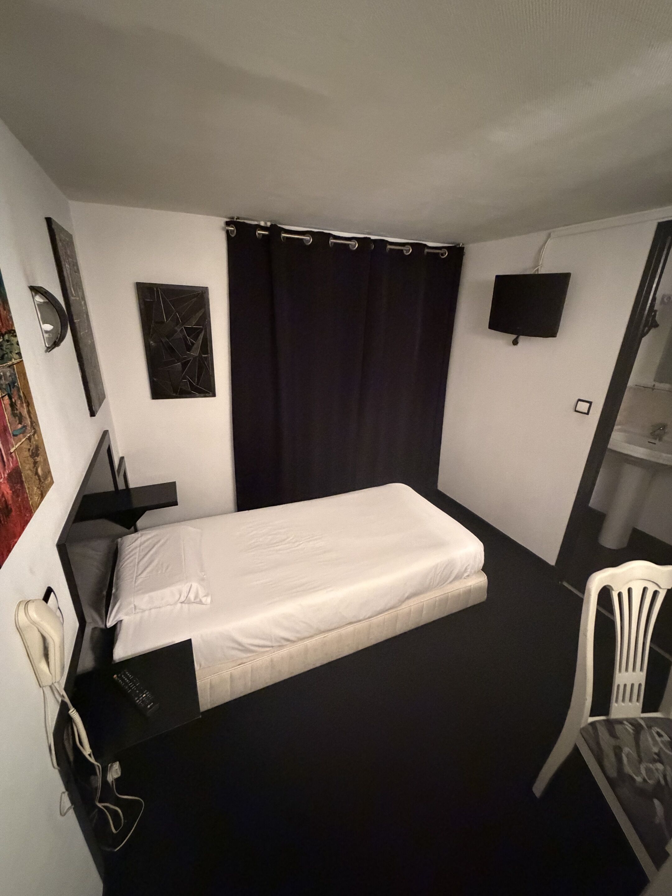 A small, single-bed room with a black curtain, a flat-screen TV, a desk with a chair, and a white chair.