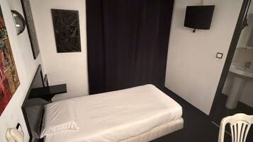 Una habitación pequeña con cama individual, cortina negra, televisor de pantalla plana, escritorio con silla y otra silla blanca.