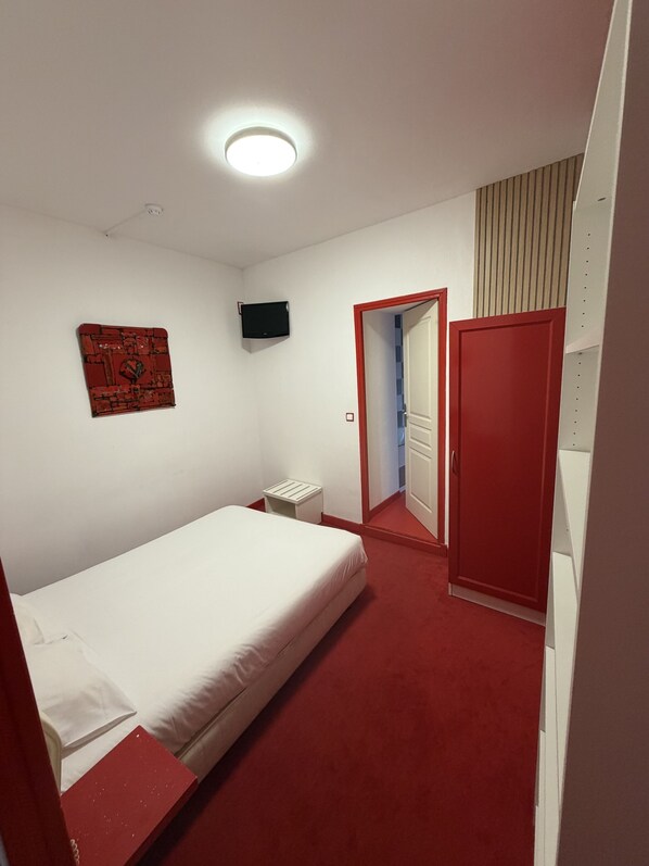 A bedroom with a single bed, a small bedside table, a red wardrobe, a television on the wall, and a door leading to another room. - Le coin des Halles (Cahors)