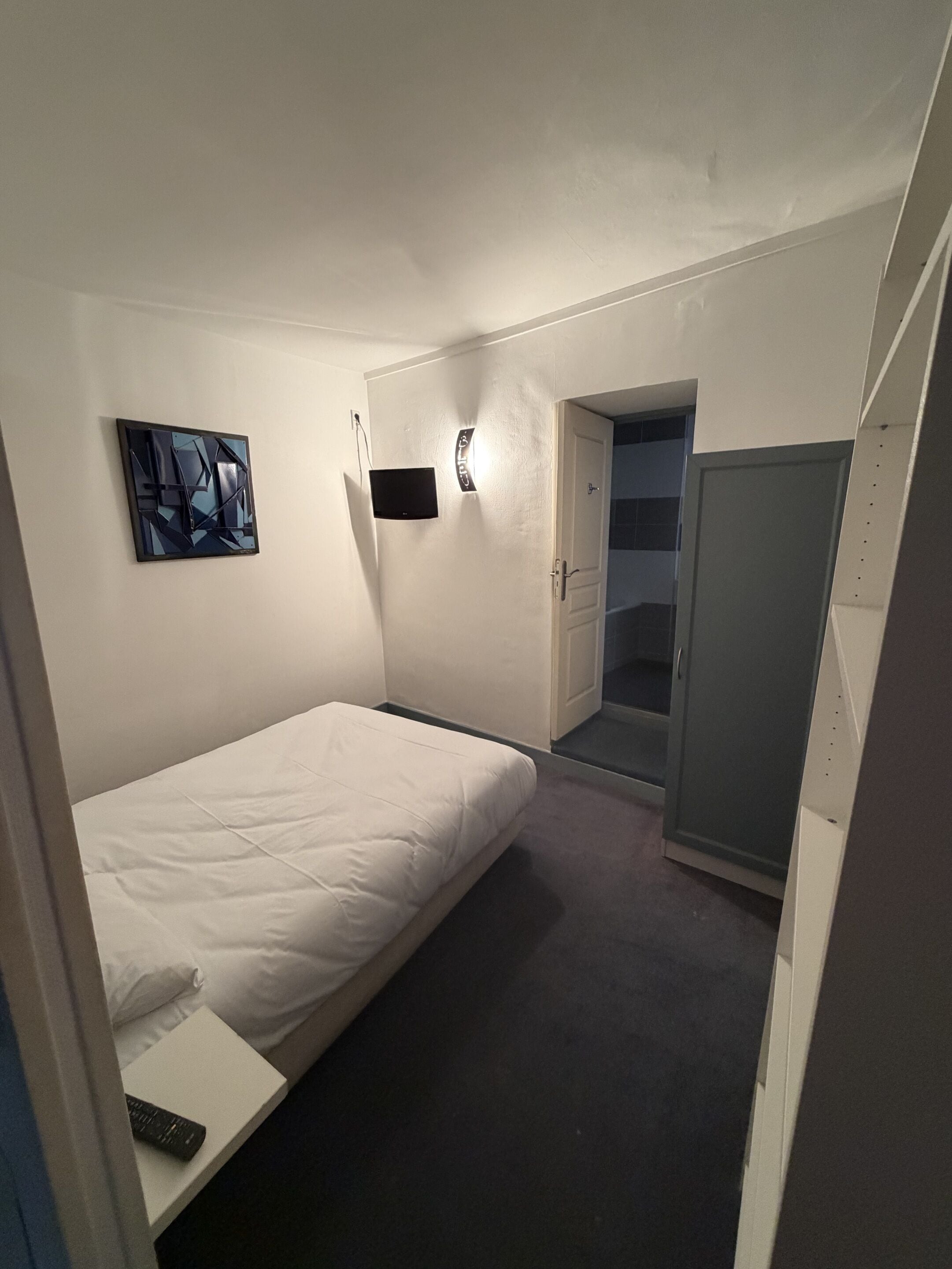 Quarto pequeno e bem iluminado, com uma cama de solteiro, mesa de cabeceira, TV fixada na parede e um armário com porta de acesso a um banheiro.