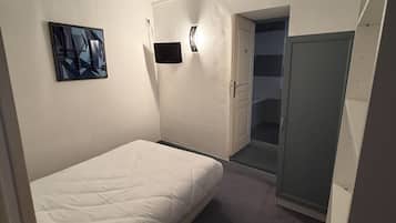 Quarto pequeno e bem iluminado, com uma cama de solteiro, mesa de cabeceira, TV fixada na parede e um armário com porta de acesso a um banheiro.