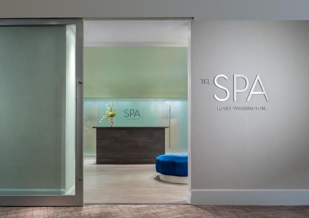 Un área de recepción con mostrador, un banco azul y una pared con el texto 'THE SPA HOTEL WASHINGTON'.