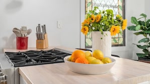 Une cuisine équipée d’une cuisinière à gaz, un saladier rempli de fruits, un vase de fleurs et une fenêtre donnant sur un beau paysage.