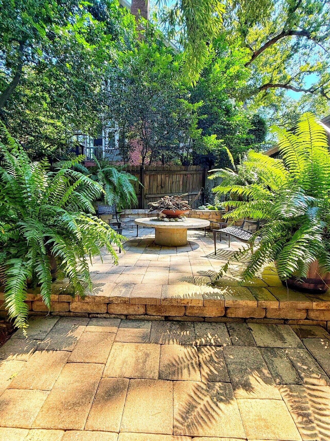 A garden patio with a stone floor, surrounded by lush greenery and a wooden fence.