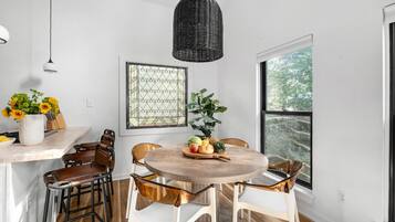 A dining area with a round wooden table, white chairs, a hanging black wicker light, and a window with a view of trees.