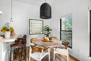 A dining area with a round wooden table, white chairs, a hanging black wicker light, and a window with a view of trees. - Inman Park Treetop Loft & Beltline (Atlanta)