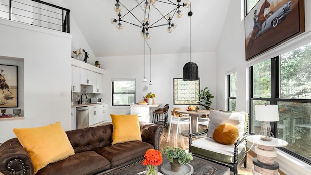 Un salon moderne avec un canapé en cuir marron, une table basse avec des livres et un vase de fleurs, un coin repas avec une table ronde et des chaises, et une cuisine en arrière-plan.