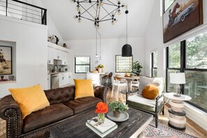 A modern living room with a brown leather sofa, a coffee table with books and a vase of flowers, a dining area with a round table and chairs, and a kitchen in the background. - Inman Park Treetop Loft & Beltline (Atlanta)