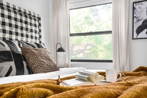 A neatly made bed with a checkered headboard, a window with a view of greenery, and a bedside table with a book and a cup.