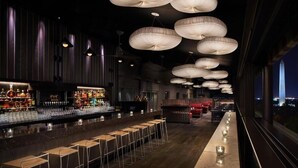 Eine Bar mit mehreren Hockern, einer Theke voller Flaschen und hängenden Lampen.