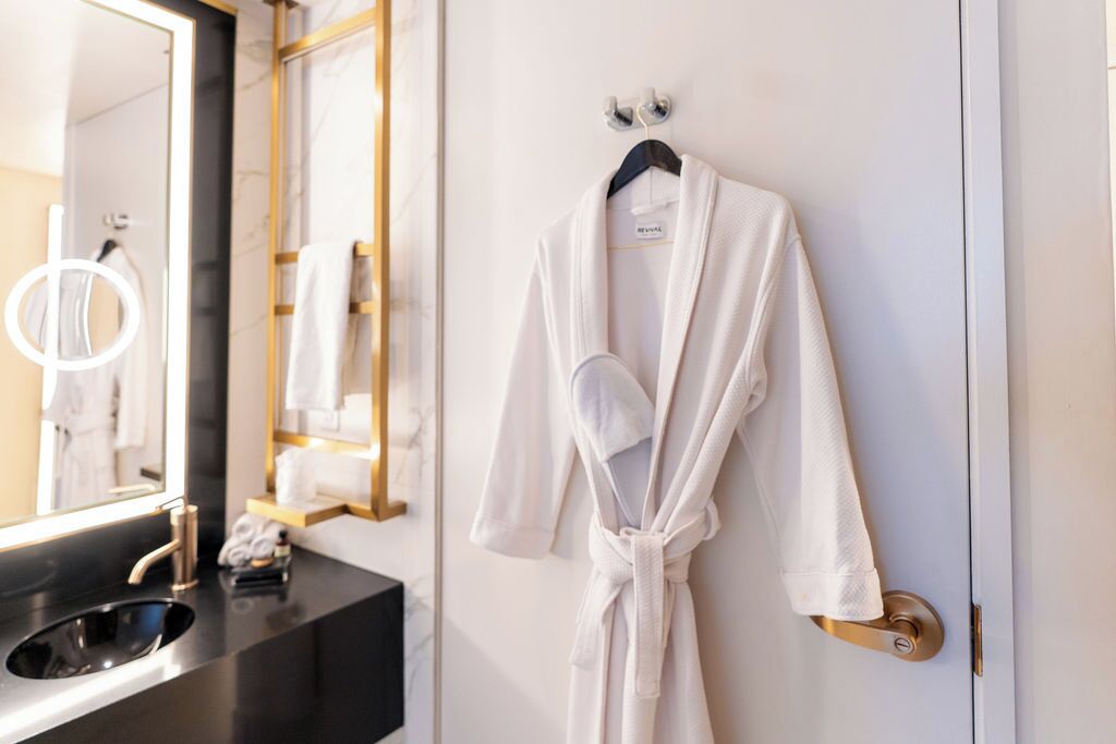 Un baño con bata blanca colgada en la puerta, un lavamanos con espejo y un perchero con toallas.