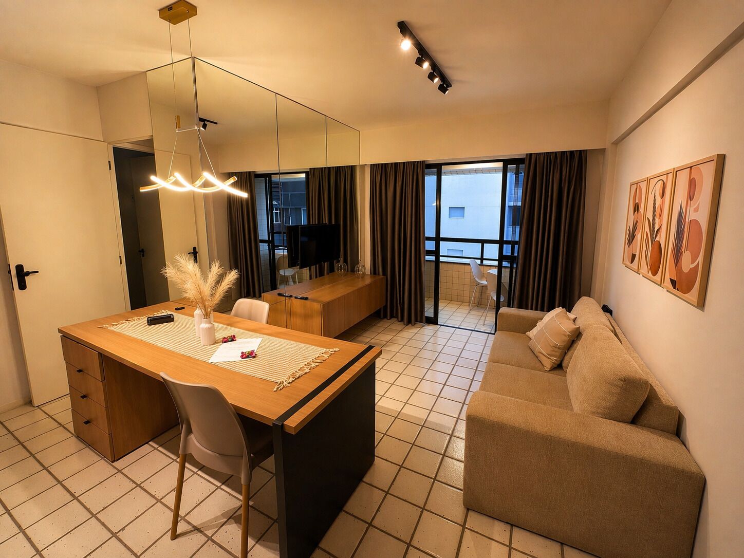 Habitación de hotel con zona de comedor, sofá, televisor y balcón con cortinas.