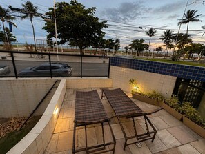 An outdoor seating area with two wicker chairs and a table, a tiled wall, and a view of the street and palm trees. - Apê Av. Boa Viagem Praia & restaurantes (Recife)