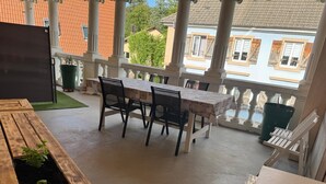 Un espace extérieur abrité, aménagé pour les repas, avec une table et des chaises, un jardinier en bois planté de végétaux, et une vue sur les bâtiments en arrière-plan.