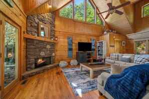 A log cabin interior with a stone fireplace, wooden walls, a ceiling fan, and a large window. - Diamond in the Rough Lakefront Cabin (Lake Lure)