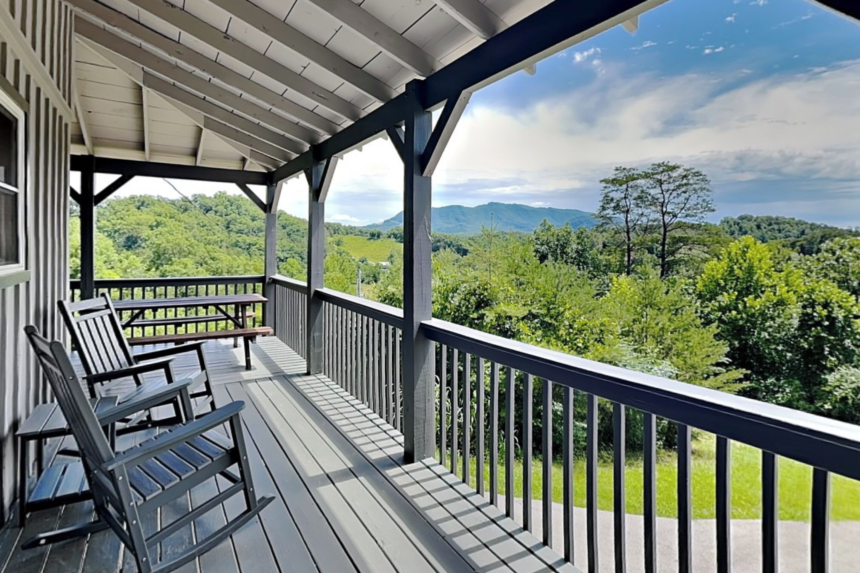 Une véranda couverte, dotée de balustrades en bois et de fauteuils Adirondack, qui offre une vue sur un paysage verdoyant et luxuriant, avec des montagnes à l’horizon.