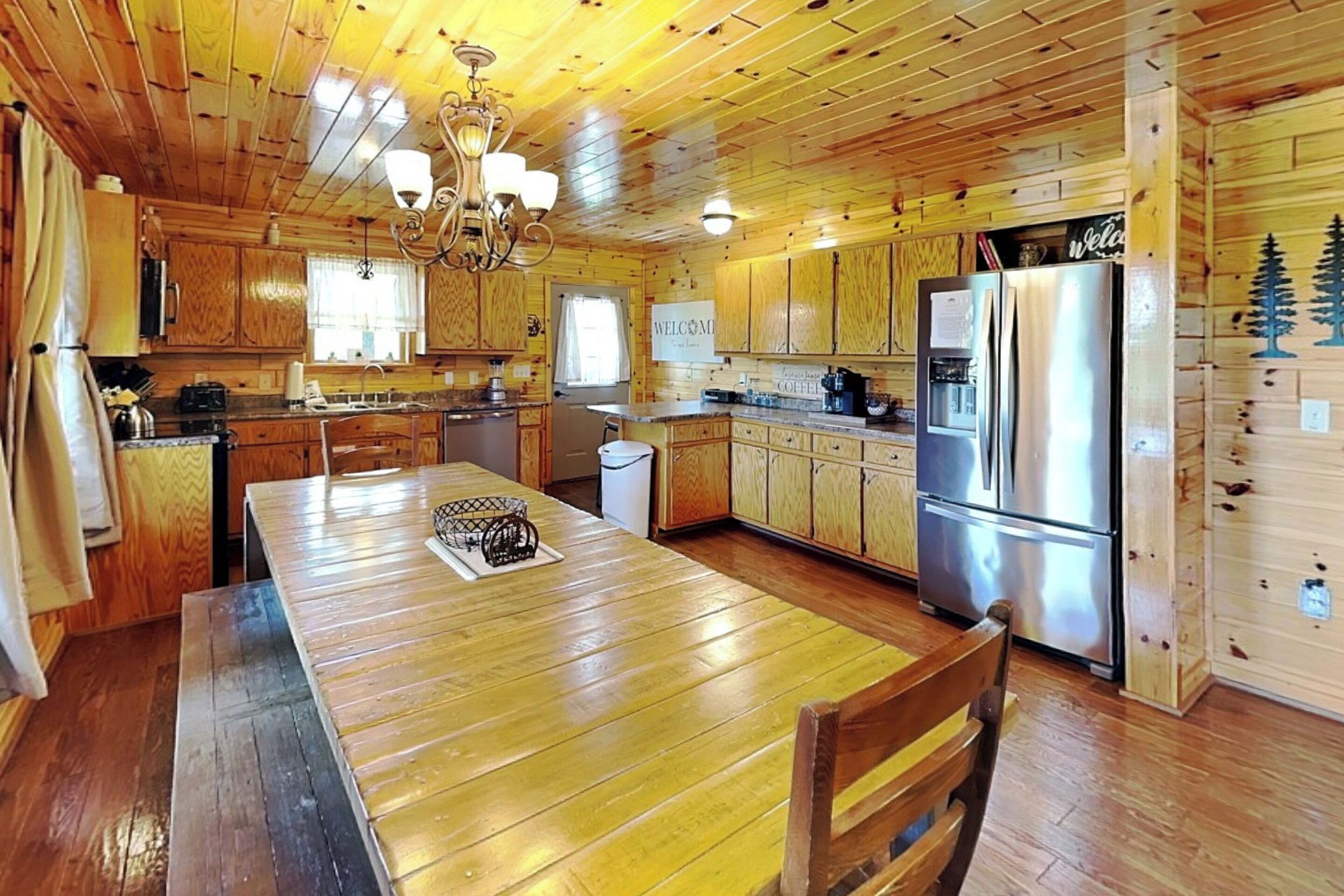 A wooden cabin interior with a central dining table, kitchen area, and a refrigerator.