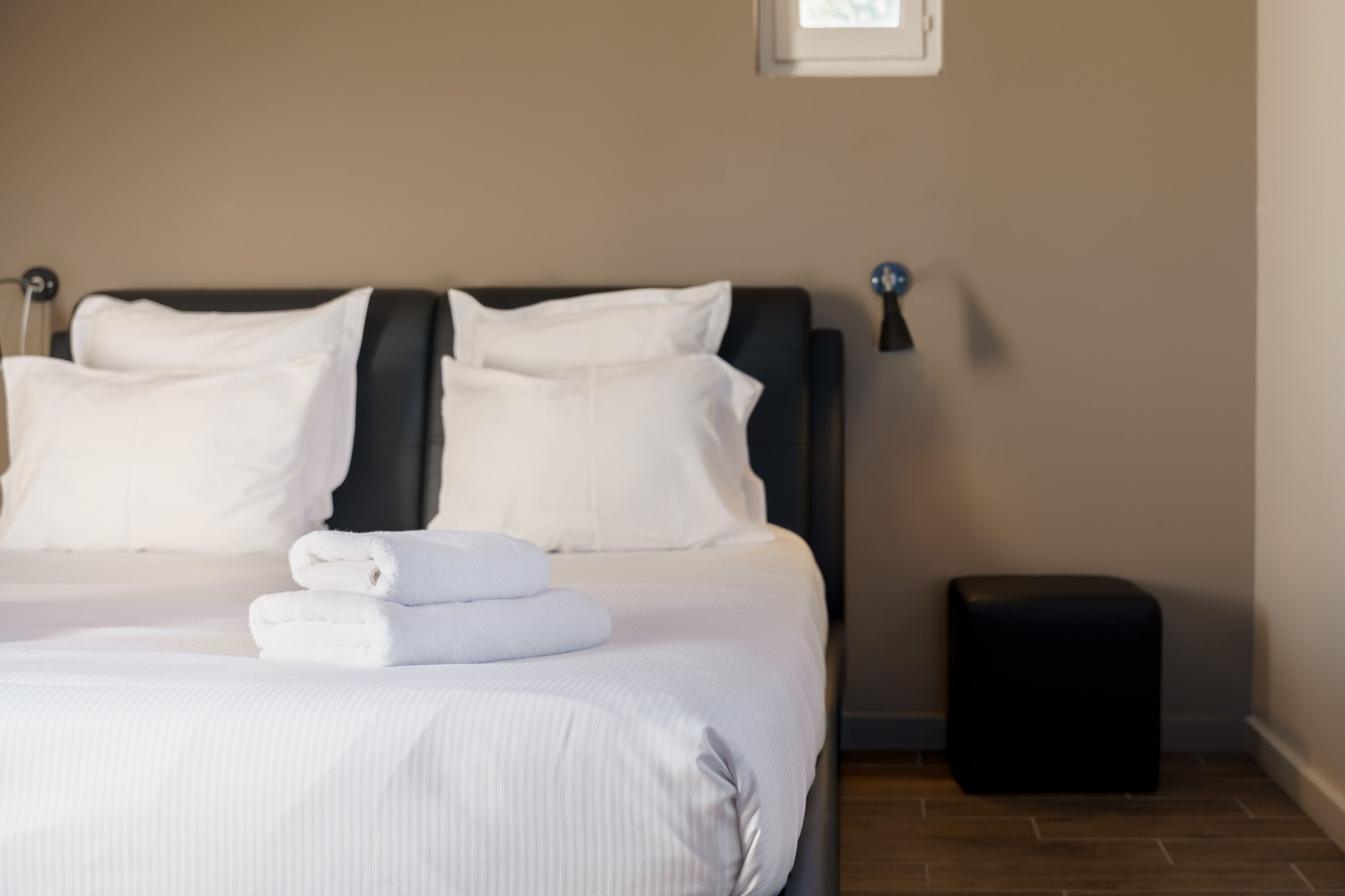 A neatly made bed with white linens and pillows, a small black ottoman, and a framed picture on the wall.