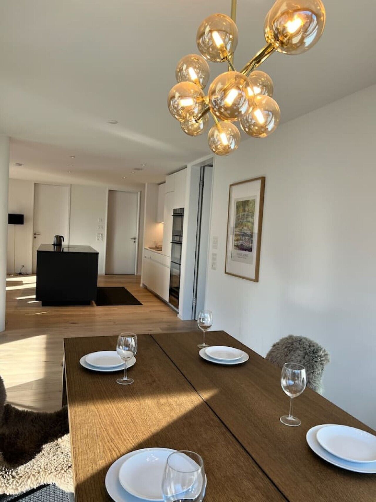 Ein moderner Essbereich mit einem Holztisch, gedeckt für vier Personen, einer Chandelüster mit kugelförmigen Glasschirm und einer Küche im Hintergrund.