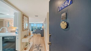 A modern apartment with a dark blue door, a kitchen with a dishwasher, a dining area with a table and chairs, and a living room with a sofa and a view of the city. - 🏝️☀️🎏 Pool, Pickle, Tennis, Beach Access all at Sun-Drenched Siesta Key Getaway⛱️🌞🐬 (Siesta Key)