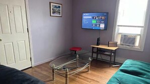 Une pièce avec une table en verre, une chaise rouge, un téléviseur fixé au mur et une fenêtre avec un climatiseur.