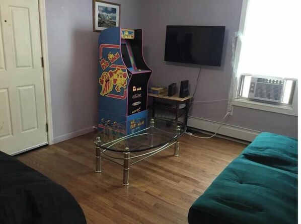 Una habitación con una mesa de vidrio, una máquina recreativa antigua, un televisor y una ventana con aire acondicionado.