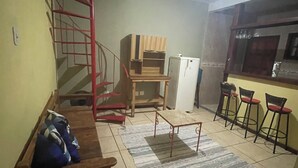 Ein kompakter Wohnraum mit einer hölzernen Bank, einem kleinen Tisch, einer roten Wendeltreppe, einer Küchenzeile mit Kühlschrank und einer Bar mit Barhocker.