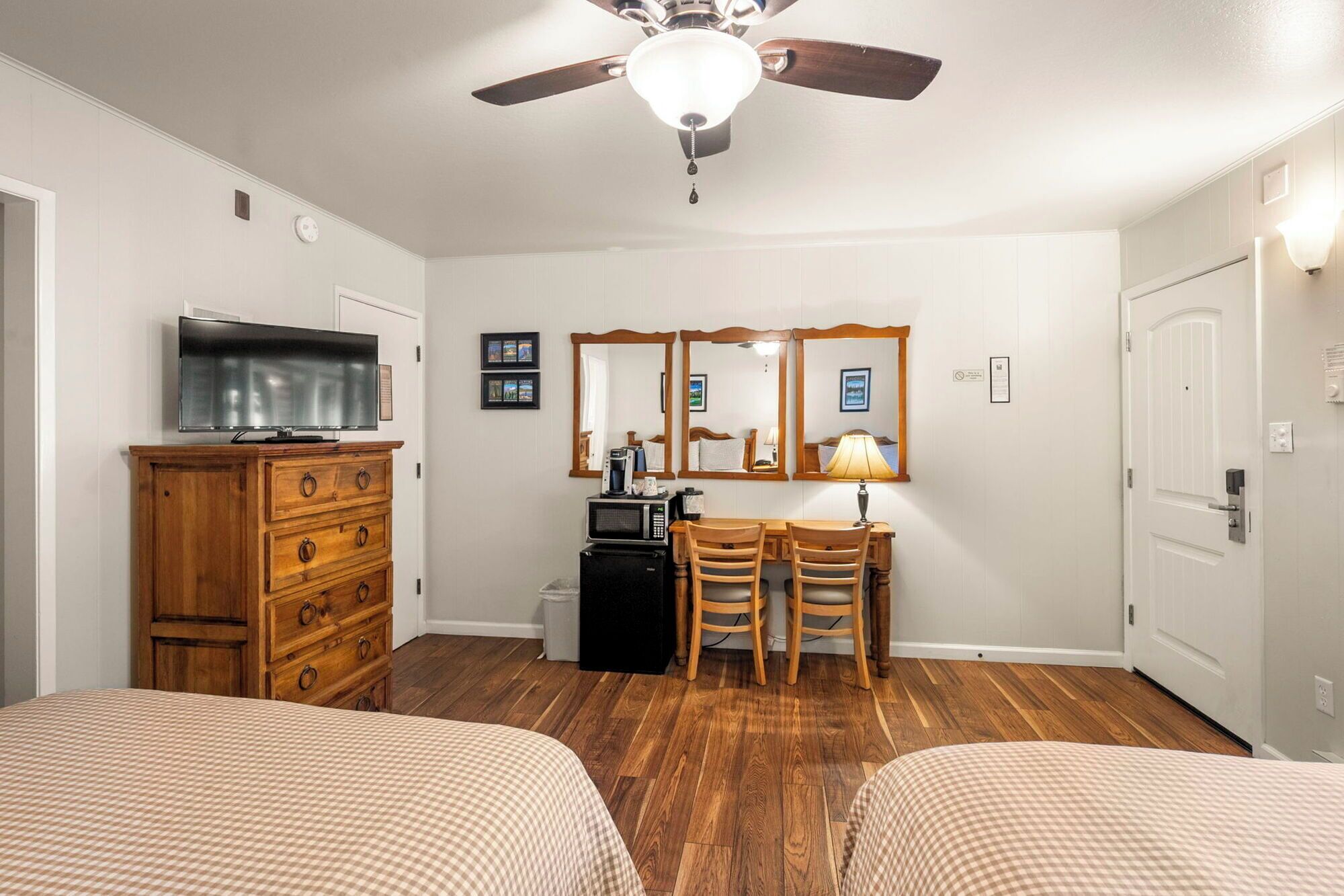 A hotel room with two beds, a wooden dresser, a desk with chairs, a ceiling fan, and a television.