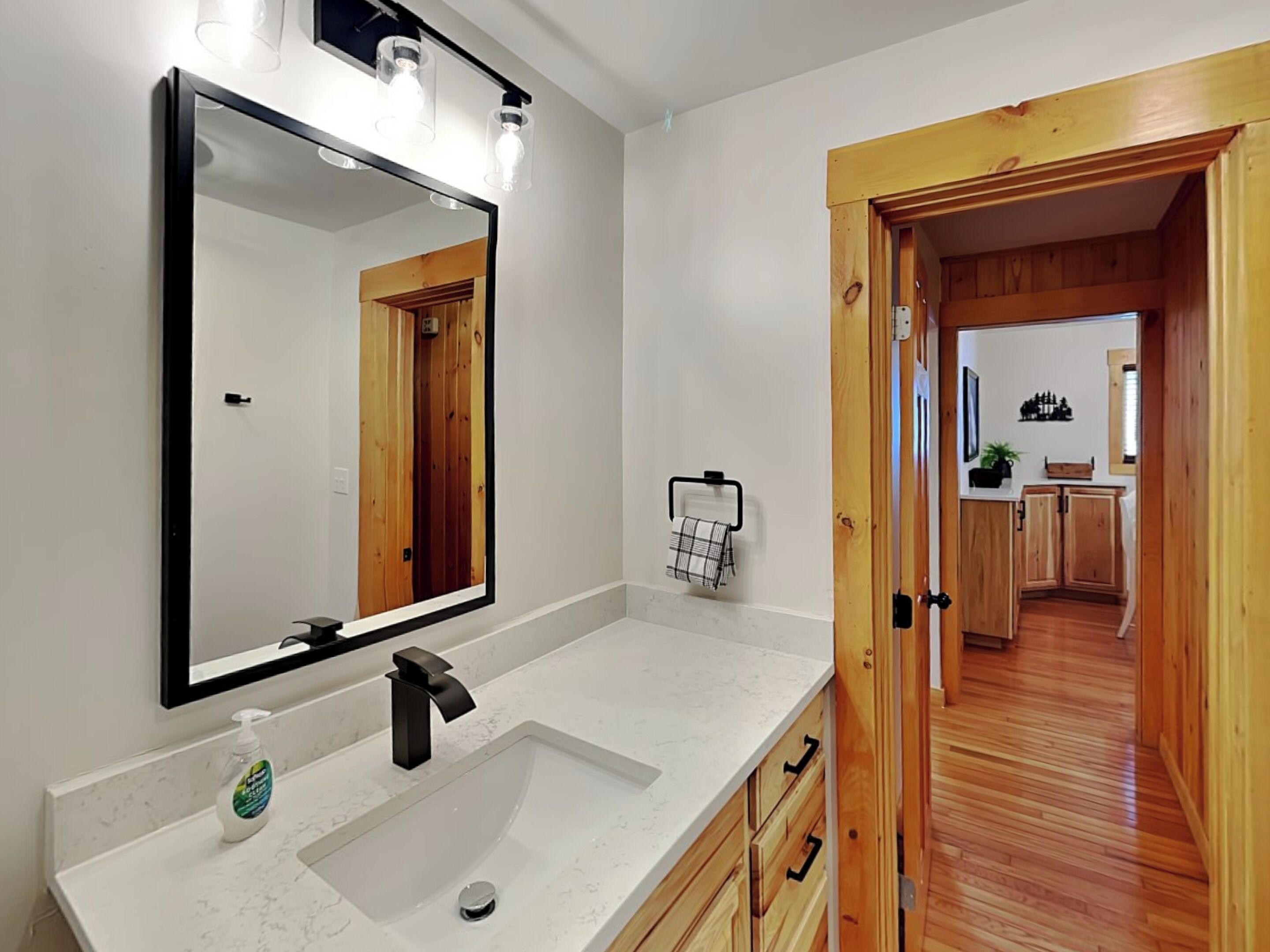 A bathroom with a large mirror, a sink with a modern faucet, and a wooden door leading to a kitchen.