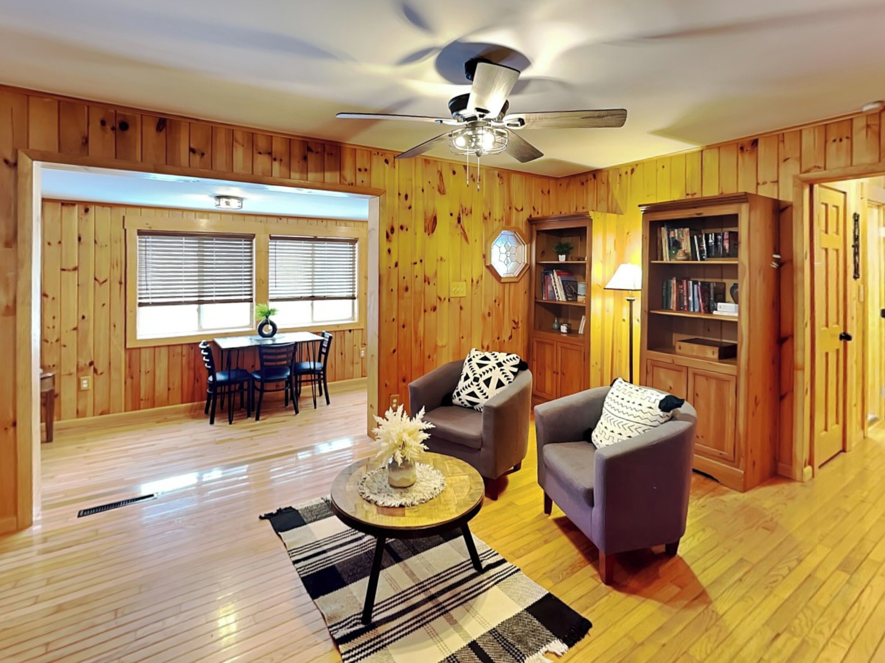 Una habitación con paneles de madera, un comedor, dos sillones, una mesa de centro, una estantería y un ventilador de techo.