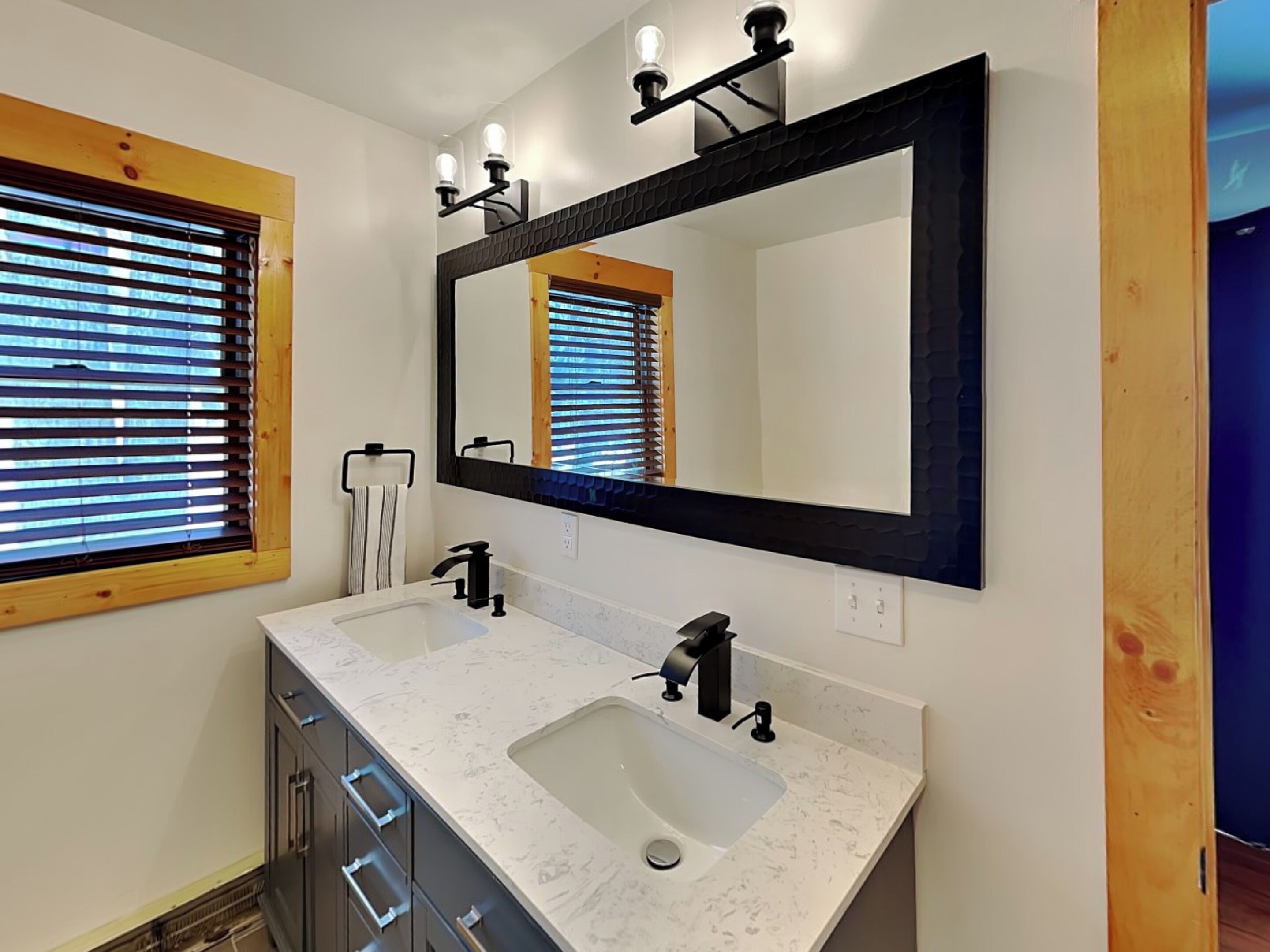 Une salle de bain avec un double lavabo, un grand miroir et des stores en bois aux fenêtres.