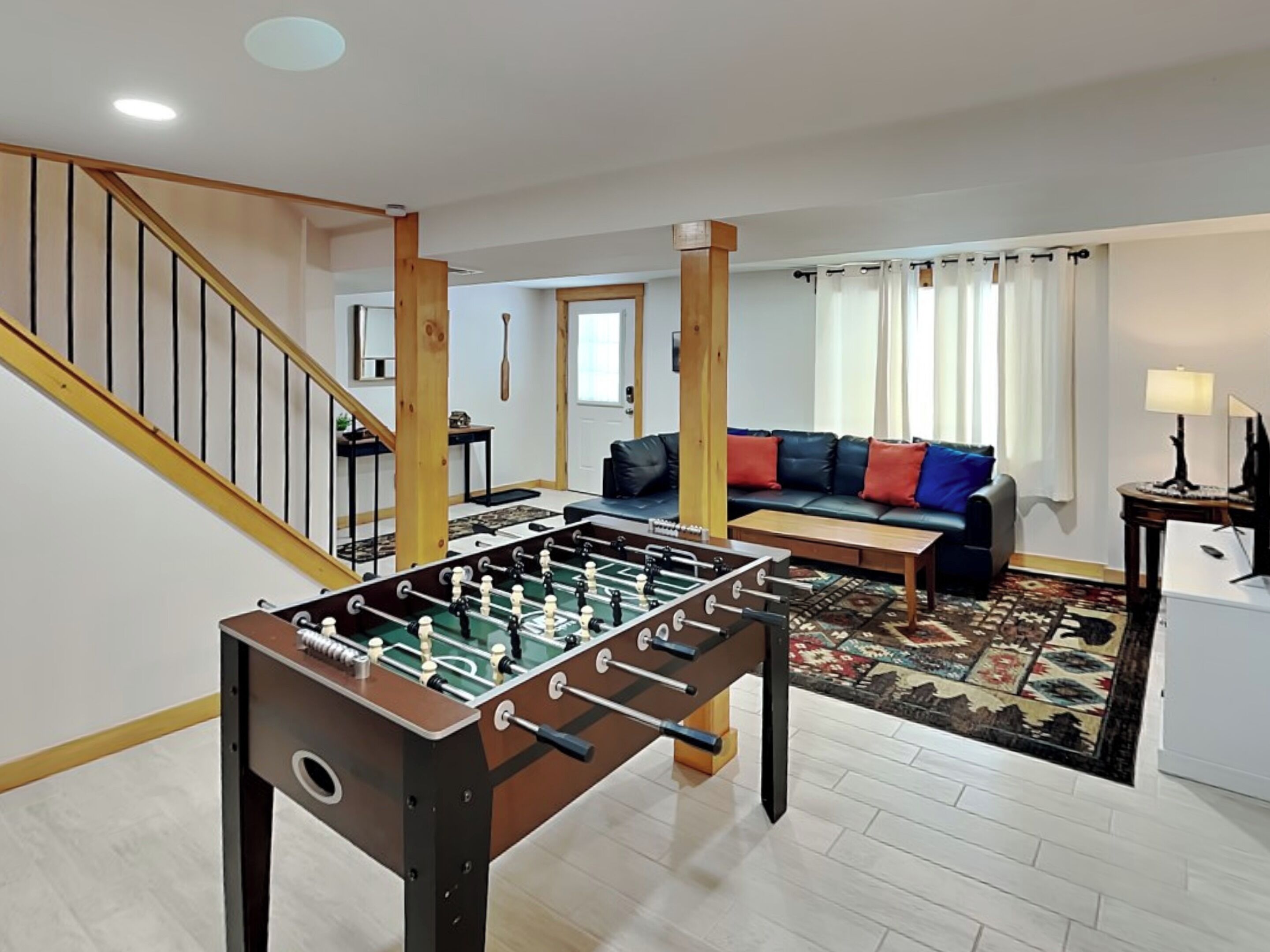 A living room with a foosball table, a sofa with colorful cushions, a patterned rug, and a staircase with wooden railings.
