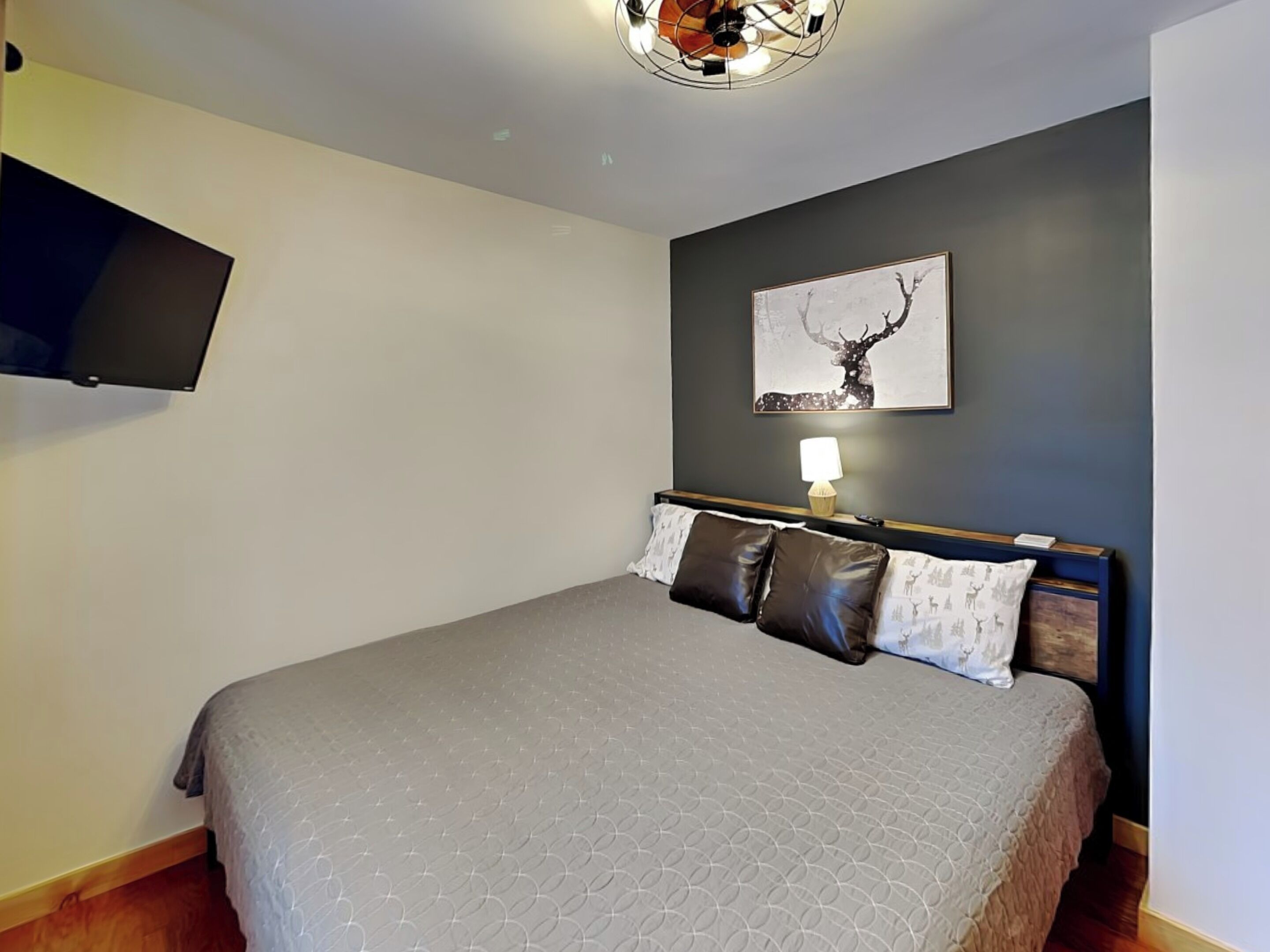 A bedroom with a bed, a mounted TV, a wall-mounted lamp, and a framed picture of a deer.