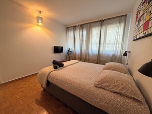A bedroom with a bed, a flat-screen TV, a wall-mounted fan, and a ceiling light. - Central 2 bedroom apartment in heart of Eaux-Vives (Genève)