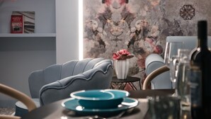 Un comedor moderno con una silla tapizada de gris, un jarrón con flores rosadas y una pared decorativa con motivos florales.