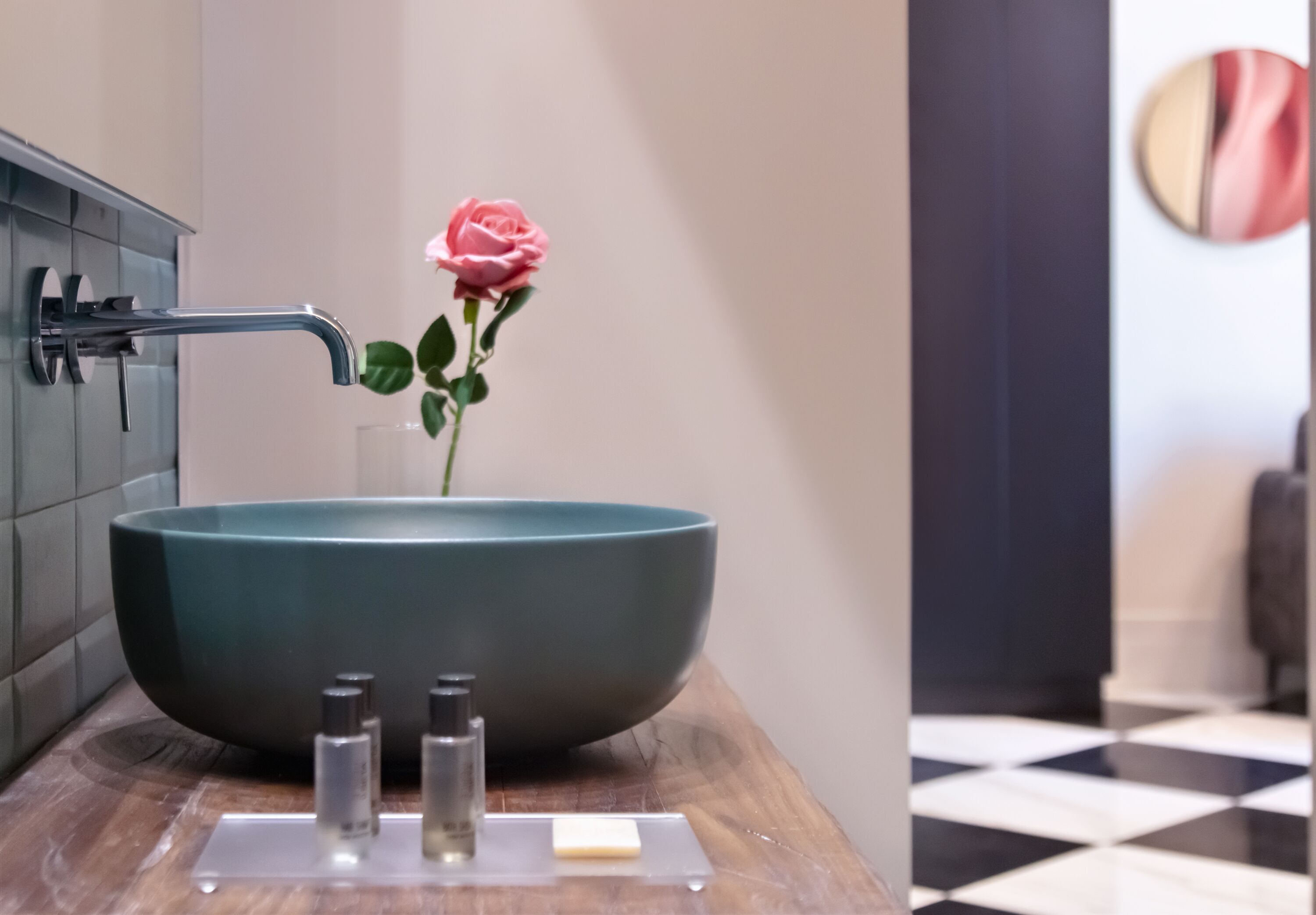 A bathroom with a large teal sink, a rose, and toiletries on a wooden countertop.
