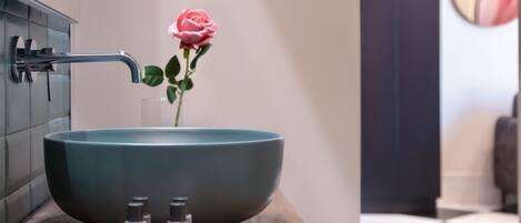 A bathroom with a large teal sink, a rose, and toiletries on a wooden countertop.