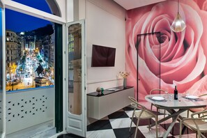 A dining area with a view of a cityscape through a window, a round table with chairs, a vase with flowers, and a wall with a large rose mural. - Lady Rose One - One Bedroom Apartment, Sleeps 4 (Naples)