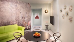 Ein modernes Wohnzimmer mit einem grünen Sofa, einem runden Tisch mit Früchten und einem großen, abstrakten Wandbild.